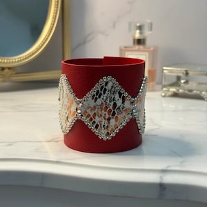 Puede incluir: Una pulsera de cuero roja con un estampado de piel de serpiente y detalles de pedrería. La pulsera presenta un diseño de banda ancha con un patrón de piel de serpiente en forma de diamante en blanco, negro y naranja. Pedrería bordea los bordes.