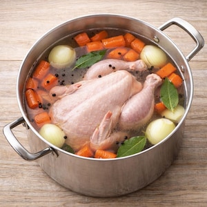 May include: A stainless steel stockpot filled with water, a whole raw chicken, carrots, onions, bay leaves, and peppercorns. The pot sits on a light brown wooden surface. The ingredients are ready to be cooked into a broth.