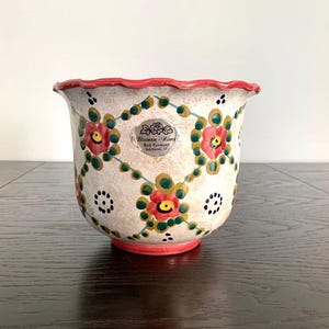 May include: A decorative ceramic planter with a scalloped red rim and base. The off-white body is adorned with red and yellow floral patterns, green vines, and black polka dots. A small sticker is visible on the front.