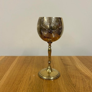 Może przedstawiać: Złoty kielich z ozdobnym wzorem na czaszy i prostą nóżką. Kielich stoi na drewnianej powierzchni.