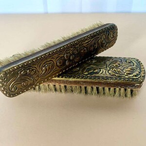 Può includere: Due spazzole per scarpe in ottone antico con decorazioni floreali ornate. Le spazzole hanno manici in legno e setole naturali.