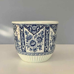 Könnte beinhalten: Ein weißer Keramik-Blumentopf mit blauem Blumenmuster. Der Topf hat eine breite, abgerundete Form mit geripptem Boden und glattem Rand. Das blaue Design zeigt Blumen und dekorative Elemente.