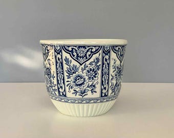 May include: A white ceramic planter with a blue floral pattern. The planter has a wide, rounded shape with a ribbed base and a smooth rim. The blue design features flowers and decorative elements.