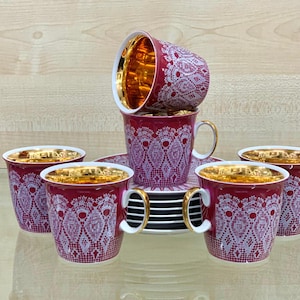 Puede incluir: Un juego de seis tazas y platillos de color burdeos con bordes y asas doradas. Las tazas y los platillos presentan un diseño de encaje blanco. El interior de las tazas es dorado. El juego está dispuesto sobre una superficie reflectante.