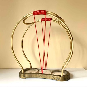 Puede incluir: Un paragüero vintage con estructura color latón y detalles rojos. El soporte presenta un diseño curvo con base rectangular y soportes verticales rojos. Diseñado para paraguas, añade un toque retro a la entrada.