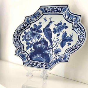 May include: A blue and white ceramic plate featuring a peacock and floral pattern. The plate has an unusual, decorative shape and is displayed on a clear stand. The design is reminiscent of Delftware.