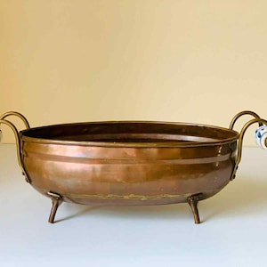 May include: An antique copper-coloured, oval bowl with three small legs and two curved handles. The handles feature white and blue ceramic detailing. The bowl is likely vintage and suitable for decorative use.