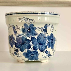 May include: A white ceramic plant pot featuring a blue floral pattern. The pot has a rounded form, a gently flared rim, and rests on three small feet. The blue flowers are painted in a detailed, stylised style.