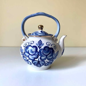 Könnte beinhalten: Weiße Keramik-Teekanne mit blauen Blumenmustern und einem blauen Griff. Der Deckel ist mit einem goldfarbenen Knauf versehen. Die Teekanne hat einen Ausguss und ist mit blauen Blumenmustern verziert.