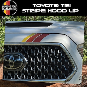 May include: White Toyota truck hood with a tri-color stripe in yellow, orange, and red. The hood features the text "TOYOTA TRI STRIPE HOOD UP" and a black honeycomb grill with the Toyota logo. The Mud & Ink Creations logo is in the upper left corner.