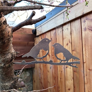 Könnte beinhalten: Metallische Silhouette von zwei Vögeln, die auf einem Ast sitzen, vor dem Hintergrund eines Holzzauns und eines Baumes mit rosa Blüten.