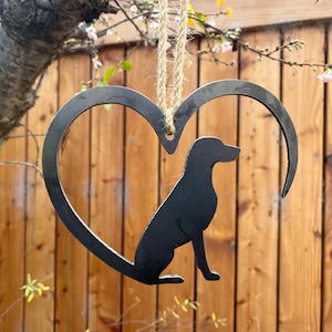 Puede incluir: Silueta negra en forma de corazón con un perro sentado en su interior, suspendida de una cuerda. El fondo es una valla de madera y ramas de árboles con flores blancas.