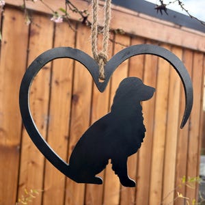 Könnte beinhalten: Schwarze Metall-Herz-Hängedekoration mit Hundesilhouette. Der Hund sitzt im Herzen. Die Dekoration wird von einem Naturfaserseil gehalten. Der Hintergrund ist ein Holzzaun.