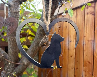 May include: A metal heart-shaped ornament with a silhouette of a dog inside. The dog is black and sits within the heart's curve. The ornament hangs from a natural fiber cord. Another heart-shaped ornament with text is visible in the background.
