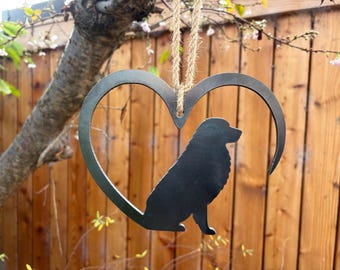 May include: A metal heart-shaped dog silhouette hanging ornament. The black dog is seated within the heart frame, suspended by a natural twine cord. The background features a wooden fence and tree branches.
