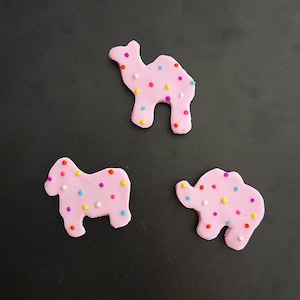 May include: Three light pink animal-shaped decorations made of a smooth material, each featuring a scattered pattern of small, colorful, circular sprinkles in shades of yellow, blue, orange, white, and purple across their surfaces.