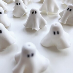 Close-up of white polymer clay ghost charms, shaped like floating sheet ghosts with unique folds and black eyes. These handmade earrings offer a cute, whimsical ghostly vibe for Halloween or quirky fashion lovers.