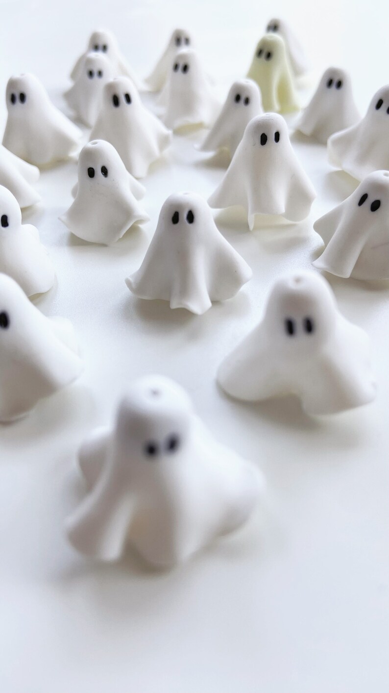 Close-up of white polymer clay ghost charms, shaped like floating sheet ghosts with unique folds and black eyes. These handmade earrings offer a cute, whimsical ghostly vibe for Halloween or quirky fashion lovers.