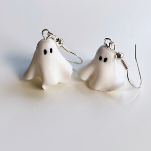 Pair of handmade polymer clay ghost earrings resembling floating sheet ghosts. Each ghost features unique organic folds and a pair of adorable black eyes, giving them a spooky yet cute look, perfect for Halloween or everyday quirky wear Silver dangle