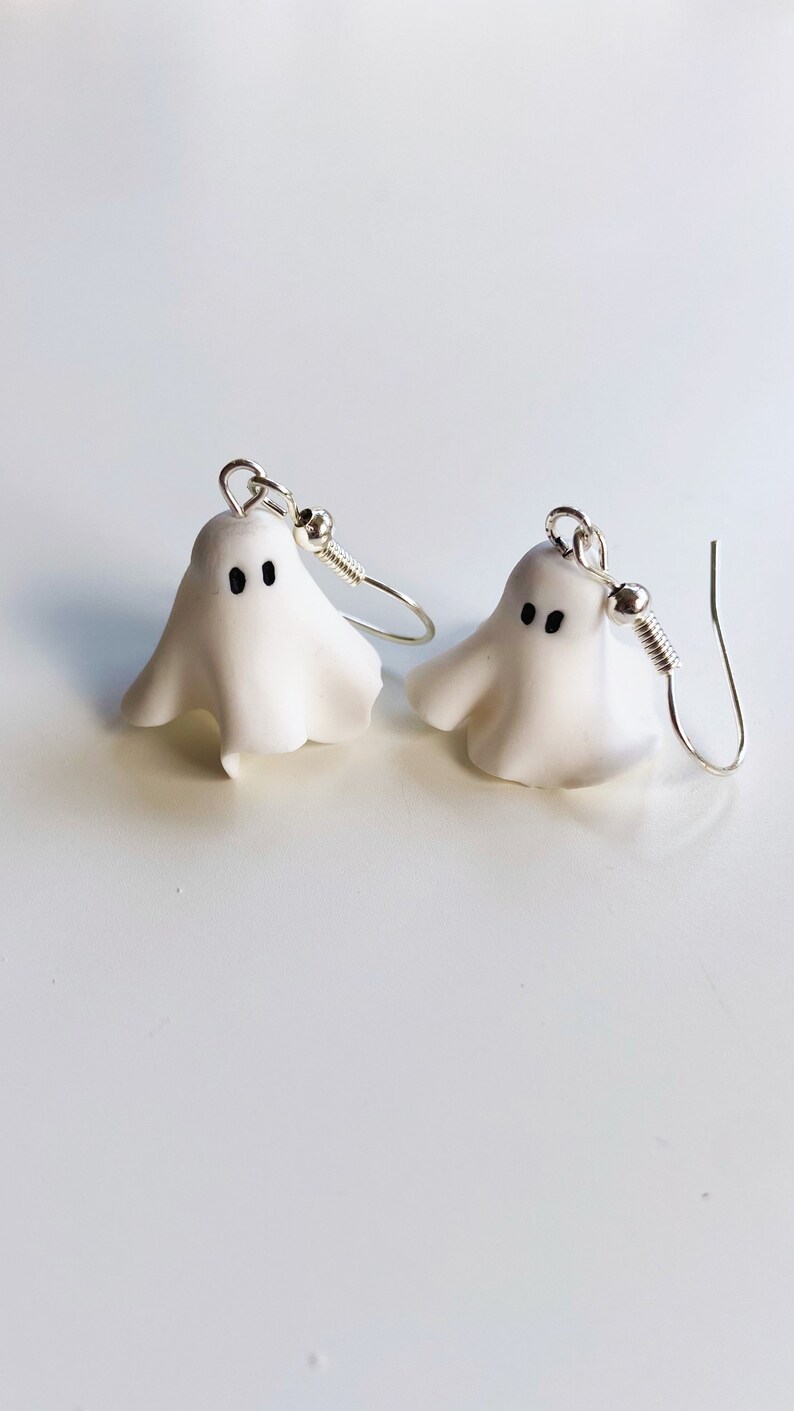 Pair of handmade polymer clay ghost earrings resembling floating sheet ghosts. Each ghost features unique organic folds and a pair of adorable black eyes, giving them a spooky yet cute look, perfect for Halloween or everyday quirky wear Silver dangle