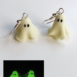 Pair of classic sheet ghost earrings, glowing in the dark. Picture shows the pair of earrings in the daylight and illuminated in a dark room, soft green scary glow. The ghost have tiny black eyes. Seem to float in the dark. Glowing classic Halloween.