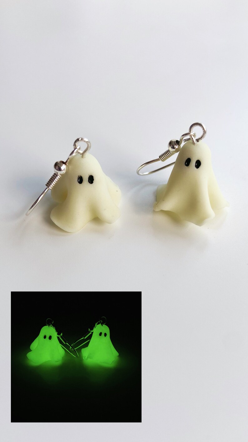 Pair of classic sheet ghost earrings, glowing in the dark. Picture shows the pair of earrings in the daylight and illuminated in a dark room, soft green scary glow. The ghost have tiny black eyes. Seem to float in the dark. Glowing classic Halloween.