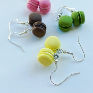 Könnte beinhalten: Ein Set mit vier Paar Ohrringen mit Miniatur-Macarons in verschiedenen Farben, darunter rosa, grün, braun und gelb. Jedes Macaron ist an einem silbernen Hakenohrring befestigt.