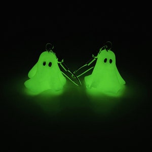 Glowing in the dark sheet ghost earrings, completely black background illuminated by the ghosts. The handmade pair of polymer clay earrings gives a green spooky glow, appearing to float. These earrings have silver dangle hooks.