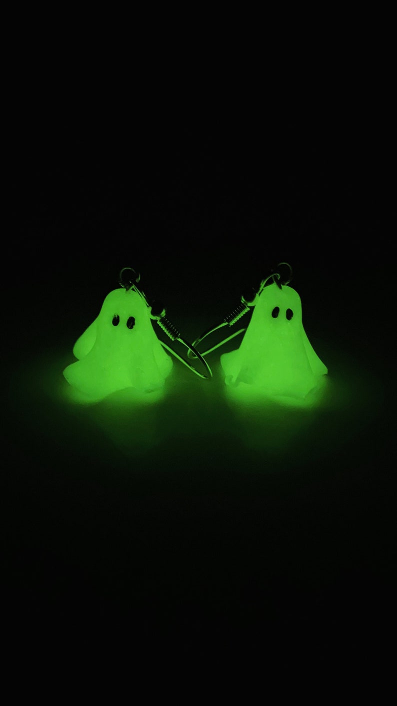 Glowing in the dark sheet ghost earrings, completely black background illuminated by the ghosts. The handmade pair of polymer clay earrings gives a green spooky glow, appearing to float. These earrings have silver dangle hooks.