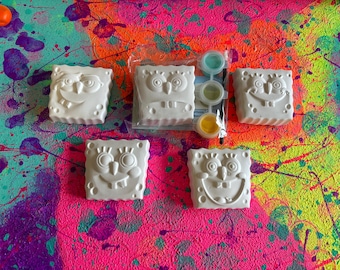 May include: Five white ceramic paint-your-own figurines of cartoon characters with different expressions. The figurines are on a colorful background with paint splatters.