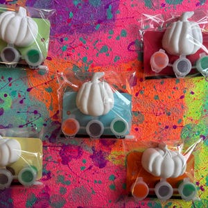 May include: Five paint-your-own pumpkin kits, each in clear packaging. Each kit contains a white pumpkin and three paint pots in orange, grey, and green. The background is a colourful splatter design in pink, orange, and teal.