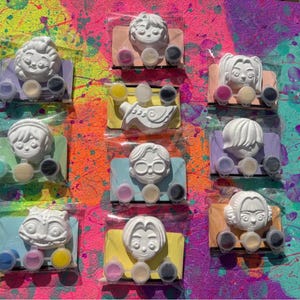 Könnte beinhalten: Neun weiße Gipsfiguren mit Malsets, jeweils in einer transparenten Plastiktüte. Die Figuren stehen auf farbigen rechteckigen Sockeln. Die Malsets enthalten kleine Töpfe mit verschiedenen Farben. Der Hintergrund ist ein lebendiges Farbspritzer-Design.