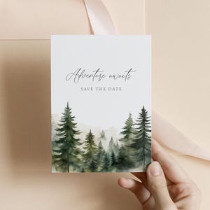 May include: A white card with the text "Adventure awaits" and "Save the date" in elegant script. The bottom of the card features a watercolor painting of a forest with green and gray trees. A hand holds the card against a neutral background.