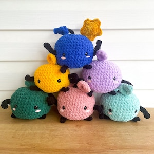 May include: A group of six crocheted plush toys in various colors, including green, yellow, pink, blue, and purple. Each toy has a round body, black eyes, and small black legs. The toys are arranged in a pyramid shape, with a yellow star on top of the blue toy.