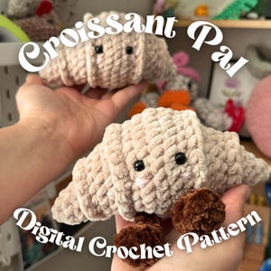 May include: Crochet croissant plush toy in a light tan color with black button eyes. The text "Croissant Pal" and "Digital Crochet Pattern" are displayed in white cursive font. The plush is held in a person's hands.