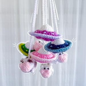 May include: A handmade mobile featuring crocheted UFOs and cow figures. The UFOs have white domes with colorful rims in pink, purple, and blue. The cows are pink and white. The mobile hangs from white cords.