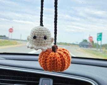 May include: A handmade car charm featuring a white crochet ghost and an orange crochet pumpkin. The ghost has black eyes and a ruffled bottom. The pumpkin has a brown stem. The charm hangs from a black braided cord.