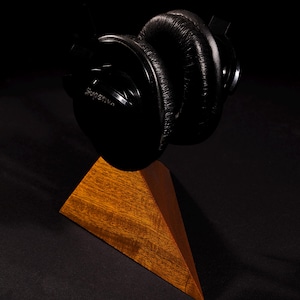 May include: Black over-ear headphones with the word "Superlux" printed on the side, resting on a wooden stand. The headphones have large, cushioned earcups and a padded headband. The stand is a geometric shape with a natural wood finish.