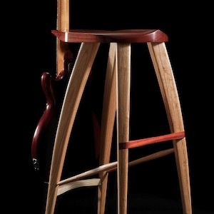 May include: A handcrafted wooden stool with a dark cherry seat and light-colored legs. The stool has a three-legged design with a red accent band. A guitar is leaning against the back of the stool.