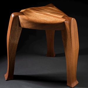 May include: A handcrafted wooden stool with a triangular seat and three curved legs. The stool is made of light brown wood with visible grain patterns. The legs have a unique, sculpted design.