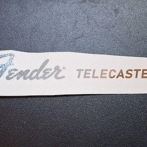 May include: Silver and gold Fender Telecaster logo sticker. The word "Fender" is in a silver script font and the word "Telecaster" is in a gold block font.