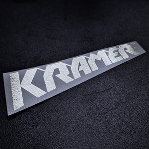 May include: A silver Kramer guitar sticker with a textured finish. The sticker is on a black background.
