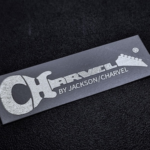 May include: A silver and black label with the text "Charvel" and "By Jackson/Charvel" with a stylized guitar graphic.