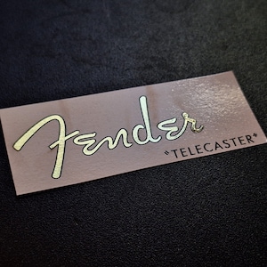 以下が含まれることがあります： 金色の筆記体で「Fender」と引用符で囲まれた「TELECASTER」という文字が書かれた長方形のステッカー。ステッカーは薄茶色の背景で、黒い質感の表面に配置されています。