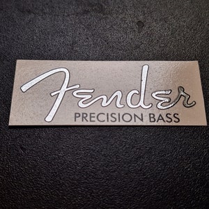 Może przedstawiać: Srebrny napis Fender Precision Bass na beżowym tle.
