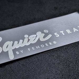 Może przedstawiać: Białe logo Squier by Fender Strat na szarym tle.