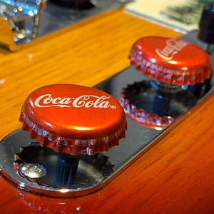 Pode incluir: Duas tampas de garrafa de Coca-Cola vermelhas são usadas como botões em uma guitarra de madeira. As tampas são brilhantes e têm o logotipo da Coca-Cola em branco.
