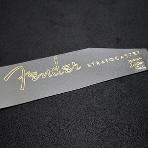 Può includere: Un adesivo dorato per la paletta della chitarra Fender Stratocaster con il testo "Original Stratocaster Parts" in un carattere più piccolo.