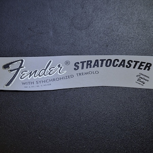 Puede incluir: Una calcomanía de guitarra Fender Stratocaster plateada y negra con el texto "Fender® Stratocaster with Synchronized Tremolo Pat. 2,741,146 3,143,028 Original Contour Body Patented".