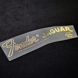以下が含まれることがあります： 「Fender」という文字が筆記体で、「JAGUAR」という文字がブロック体で書かれた、金色のフェンダー・ジャガー・ギターのデカール。デカールには、「WITH SYNCHRONIZED FLOATING TREMOLO」と「OFFSET CONTOUR BODY」という文字も書かれている。
